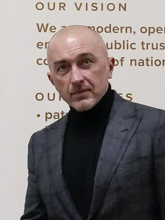 Лисий чоловік у сірому піджаку та чорній водолазці на тлі стіни з написом Our Vision